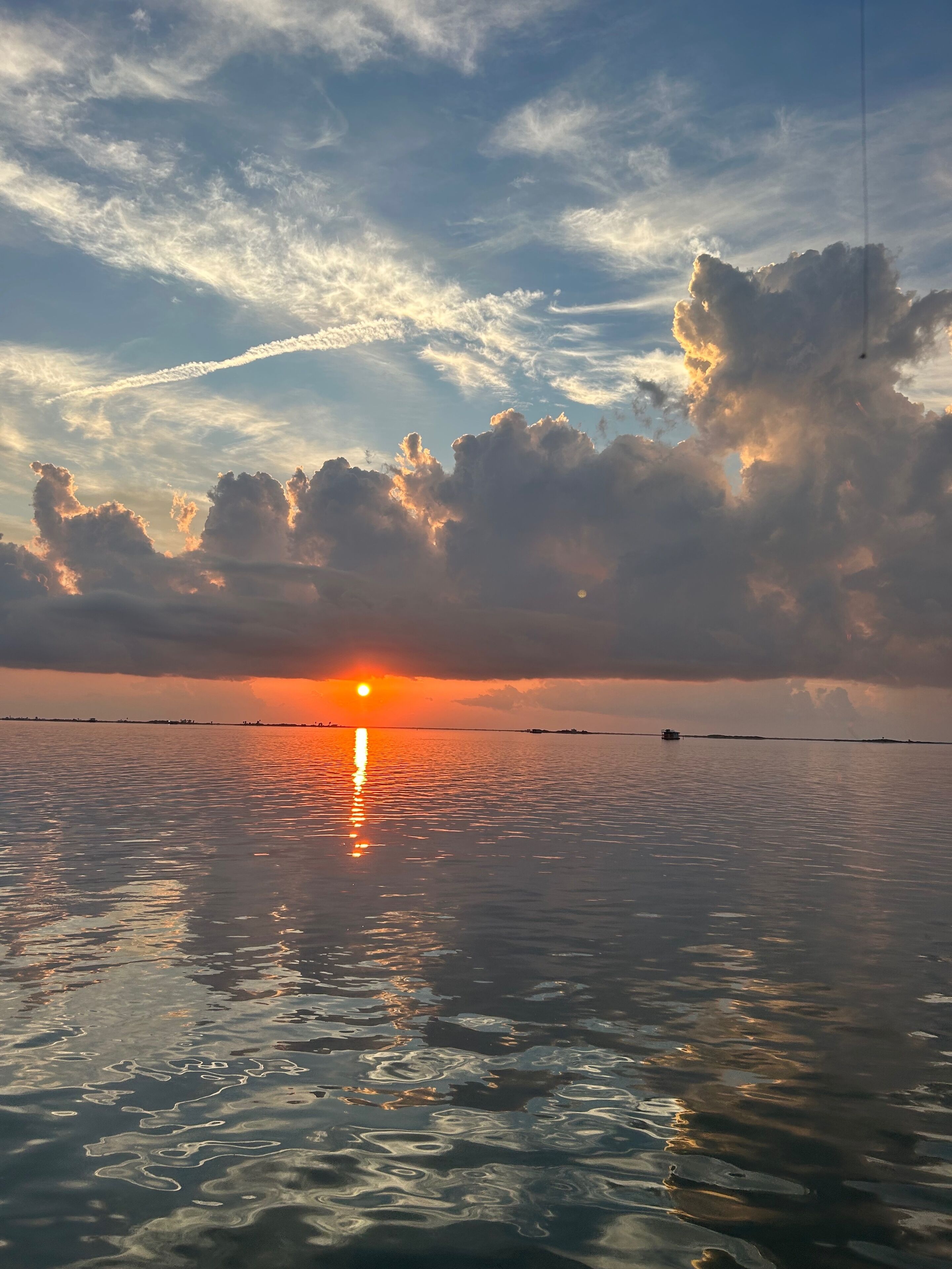A sunset over a calm body of water with boats, clear skies, and a visible contrail.