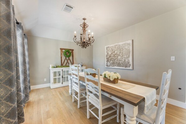 A dining room with a wooden table, white chairs, a chandelier, and a framed artwork on the wall.