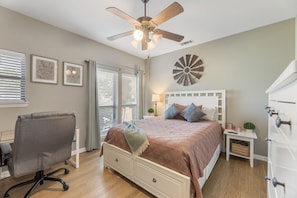A bedroom with a bed, a desk, a chair, a ceiling fan, and a wall clock.