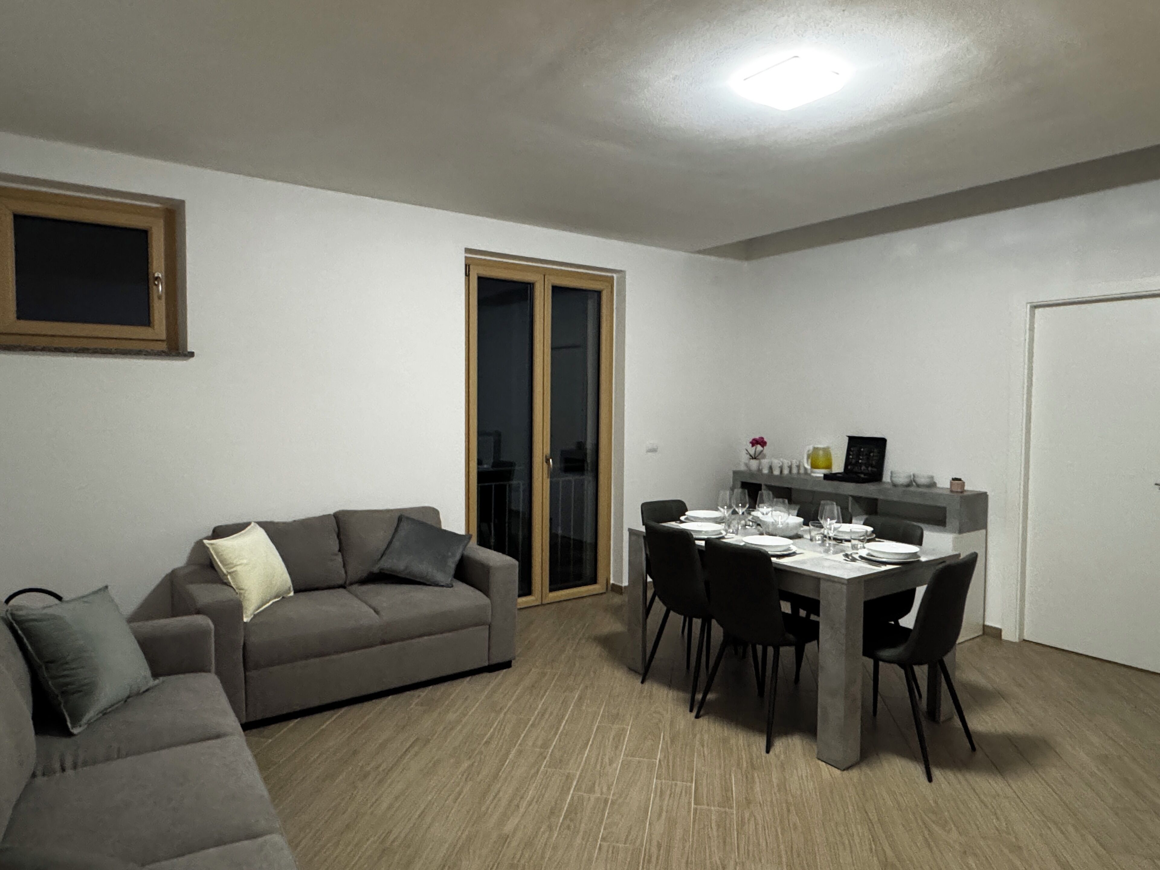 A dining area with a table set for four, a grey sofa, and a sliding glass door leading to an outdoor area.