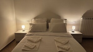 Ein ordentlich bezogenes Bett mit zwei Kissen, zwei Nachttischlampen und gefalteten Handtüchern auf Holzboden.