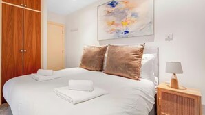 Un letto rifatto con lenzuola bianche e due cuscini, un armadio in legno, un quadro appeso al muro e un comodino con una lampada.