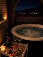 A hot tub with a city view at night, a candle, a bottle of wine, and a tray with food and drinks.