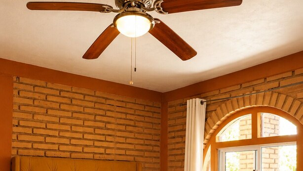 Un dormitorio con cama, cabecera de madera, ventilador de techo, ventana con cortinas y pared de ladrillo.