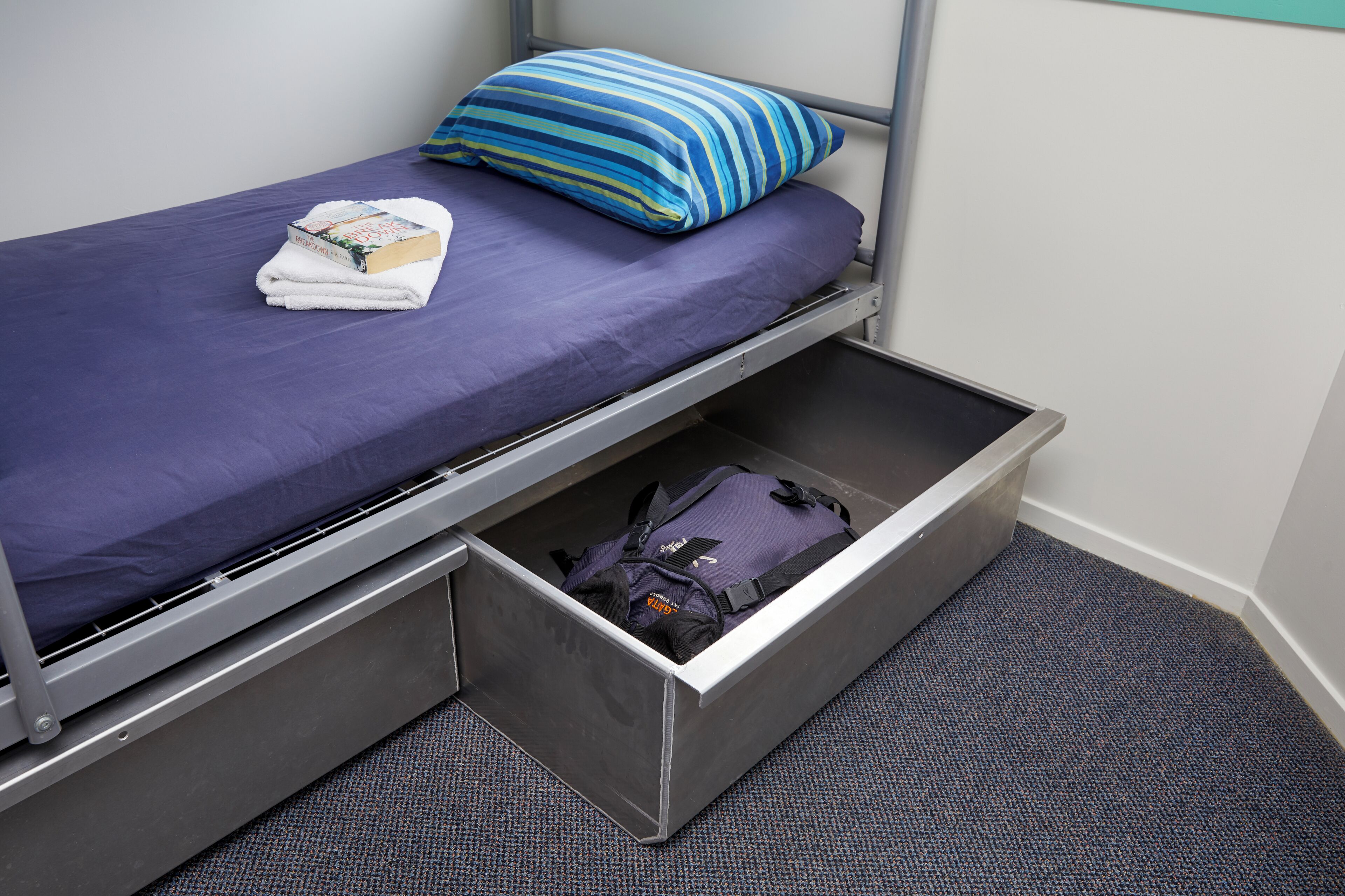 A single bed with a blue mattress and a striped pillow, a folded towel, and a backpack in a storage drawer.