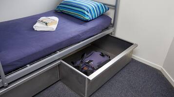 A single bed with a blue mattress and a striped pillow, a folded towel, and a backpack in a storage drawer.