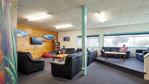 A spacious lounge area with multiple seating options, including sofas and chairs, a coffee table, and a television on the wall. - Pacific Coast Lodge and Backpackers (Tauranga)