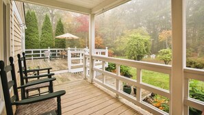 Eine überdachte Veranda mit Holzboden, weißen Geländern und Blick auf einen Garten mit Bäumen und Sträuchern.