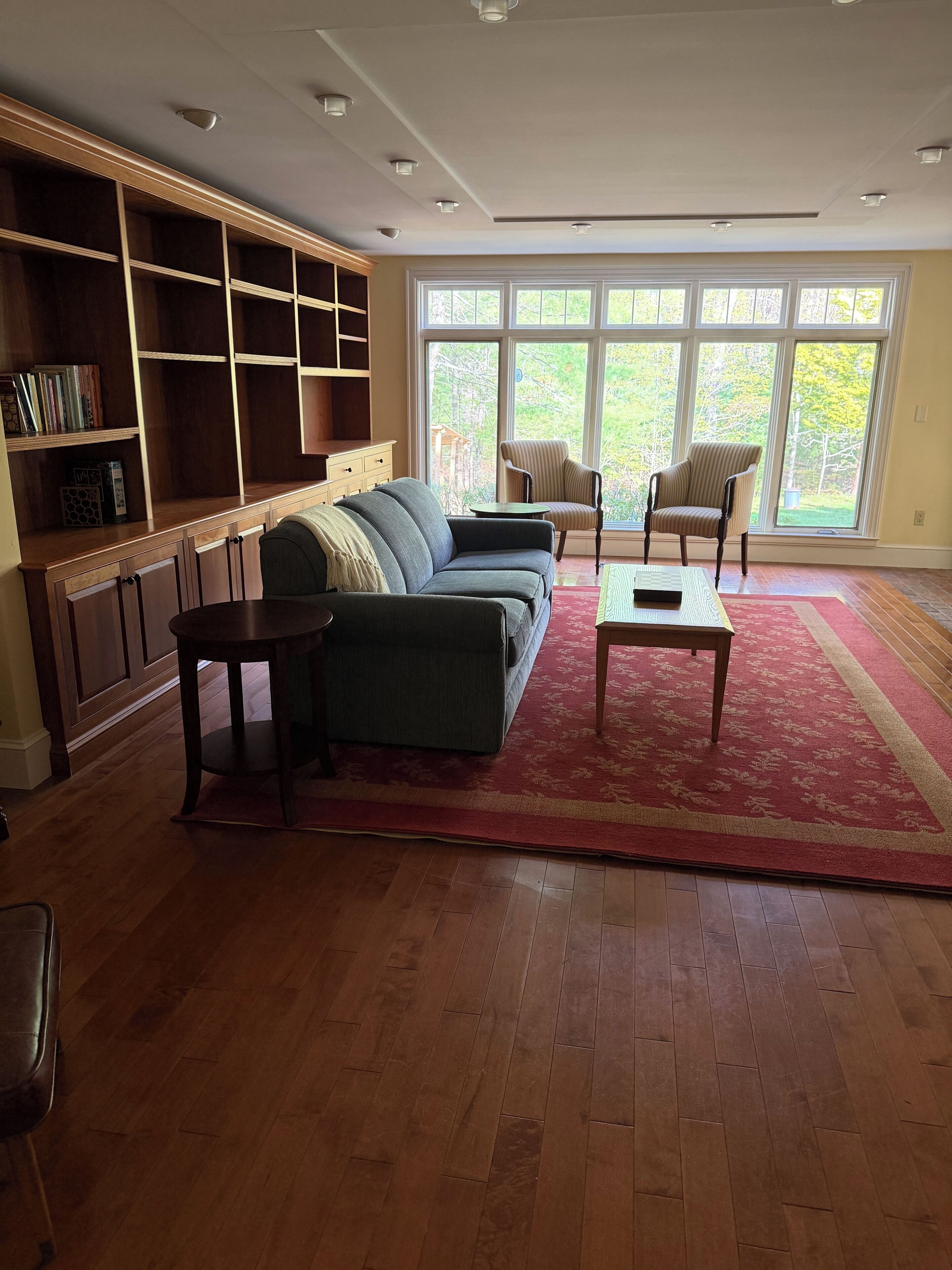 Ein Wohnzimmer mit einer Couch, Sesseln, einem Couchtisch, einem Bücherregal und großen Fenstern.