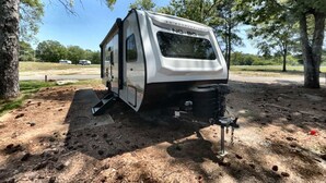 Una caravana blanca estacionada en un camping, con árboles y un cielo despejado al fondo.