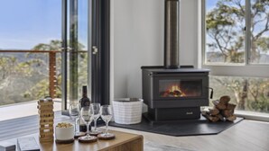 A cozy room with a wood-burning stove, a table with wine glasses and a bottle, and a view of trees through large windows. - Blue Mountains Views (WENTWORTH FALLS)