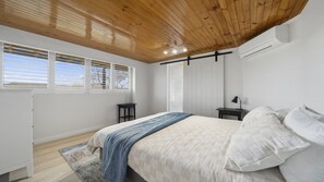A bedroom with a wooden ceiling, white walls, a bed with white and blue bedding, two nightstands, a desk, and a window with blinds. - Blue Mountains Views (WENTWORTH FALLS)