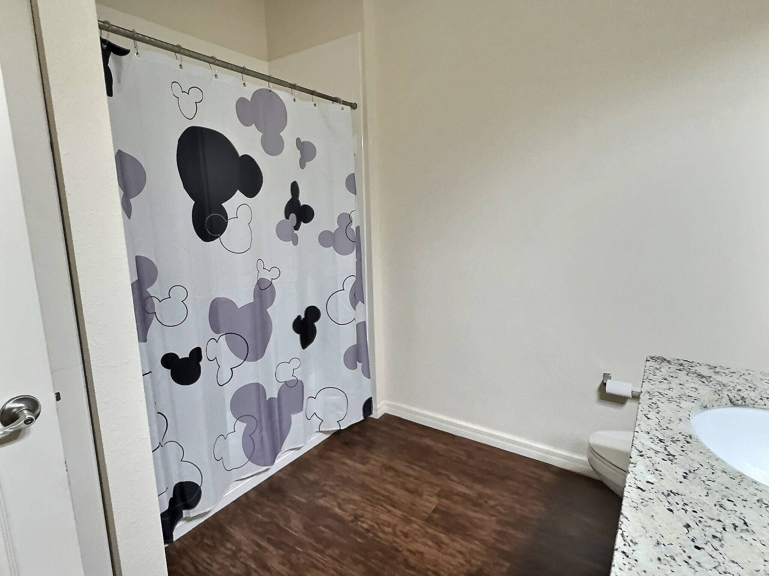 Et badeværelse med bruseforhæng med Mickey Mouse-mønster, en hvid vask med marmoreret bordplade, og trægulv.