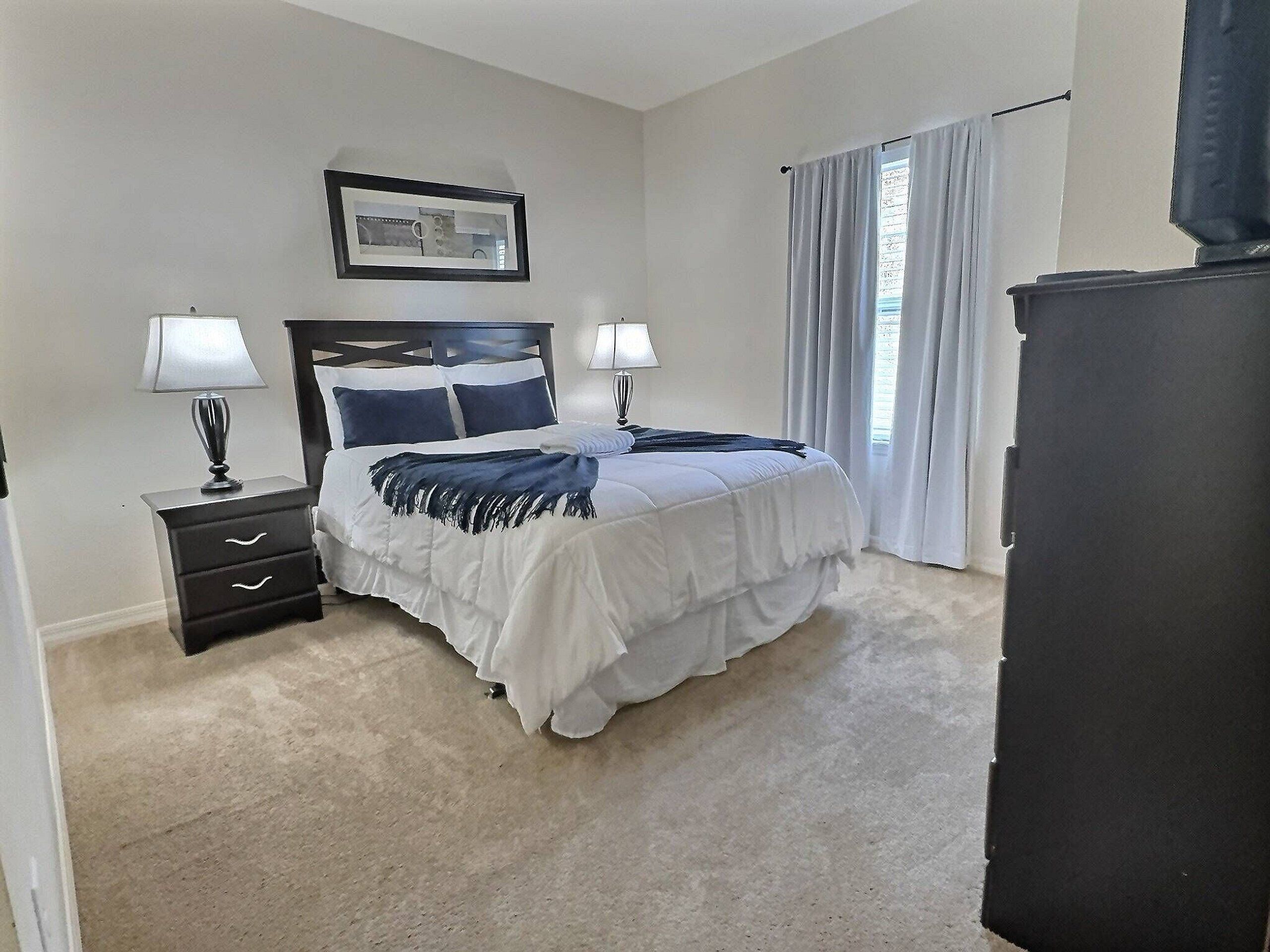 A bedroom with a bed, two bedside tables, a nightstand, a framed picture on the wall, and a window with curtains.