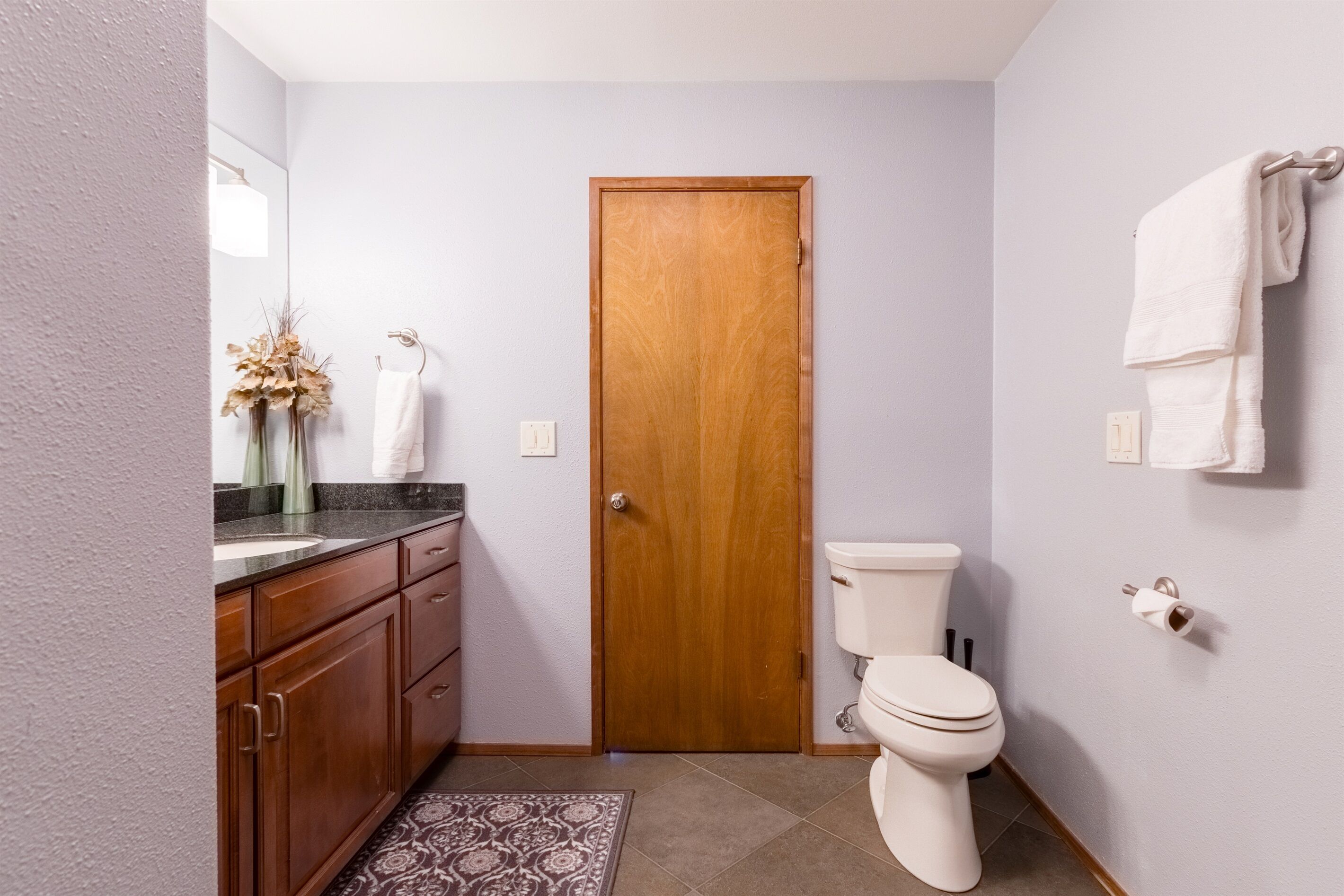 Une petite salle de bain dotée d’une porte en bois, de toilettes, d’un lavabo avec un plan de toilette en marbre et d’un tapis à motifs.