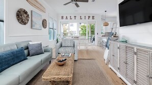 A spacious living room with a grey sofa, a woven coffee table, and a ceiling fan. A kitchen area with white cabinets and a TV is visible in the background. - Luxury 4BR/4.5BA Waterfront Home in Sandestin • Golf Carts/Golf • Pool • Beach (Miramar Beach)