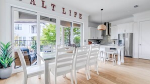 A modern kitchen with white cabinetry, a stainless steel refrigerator, and a dining area with white chairs and a glass table. - Luxury 4BR/4.5BA Waterfront Home in Sandestin • Golf Carts/Golf • Pool • Beach (Miramar Beach)