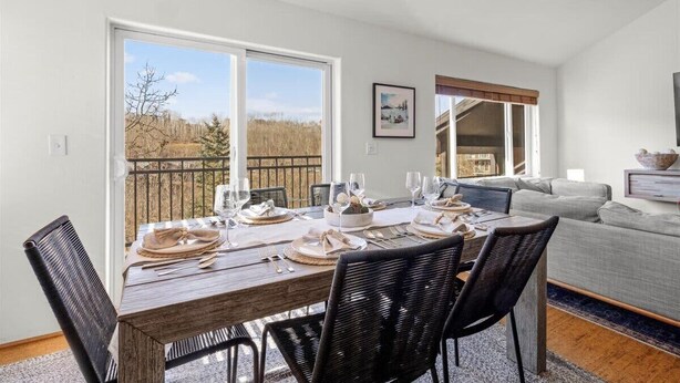 Un coin repas avec une table en bois dressée pour quatre personnes, des chaises noires et une vue sur une forêt, visible à travers de grandes fenêtres.