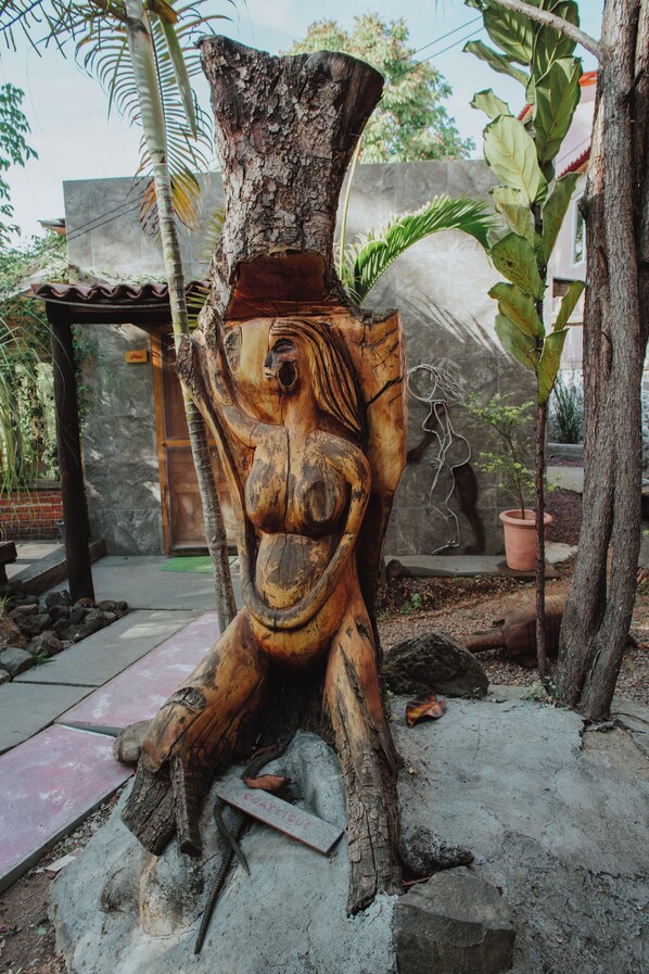 Escultura de fusta d'un rostre somrient amb els braços estesos, situada en un jardí amb una paret de pedra i plantes en testos al fons.