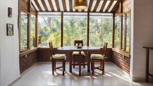 Un espace repas avec une table ronde et des chaises, de grandes fenêtres donnant sur un paysage verdoyant et un plafond en bois.