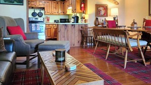Un salon chaleureux avec un canapé, un pouf et une table basse, un coin repas avec des chaises et une table, et une cuisine dotée de meubles en bois et d’appareils électroménagers.