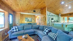 Interior acolhedor de cabana com um amplo sofá em formato de L, teto de madeira e área de cozinha com armários verde-claros.