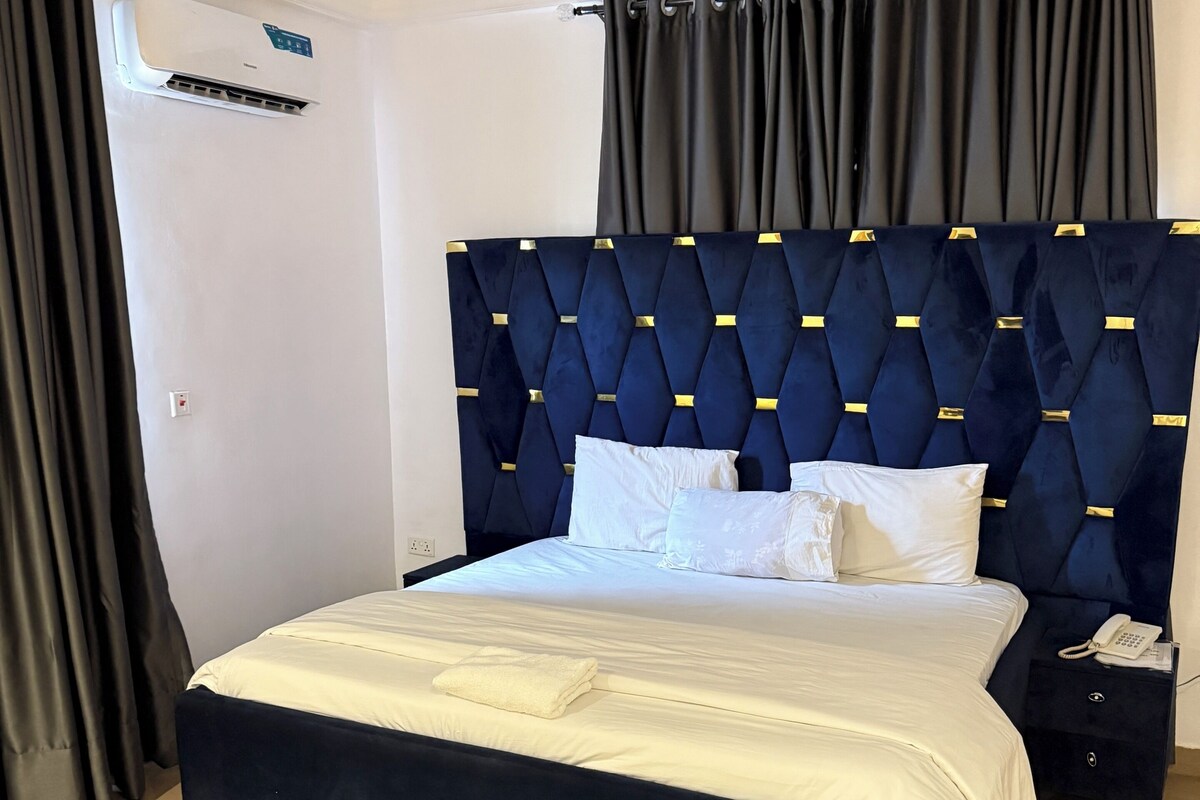 Una cama bien hecha con un cabecero oscuro, una mesita de noche con un teléfono y una alfombra estampada sobre un suelo de baldosas.
