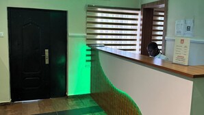 Een receptiegedeelte met een receptiebalie, een zwarte deur, een raam met zonwering en een airconditioning op de muur.