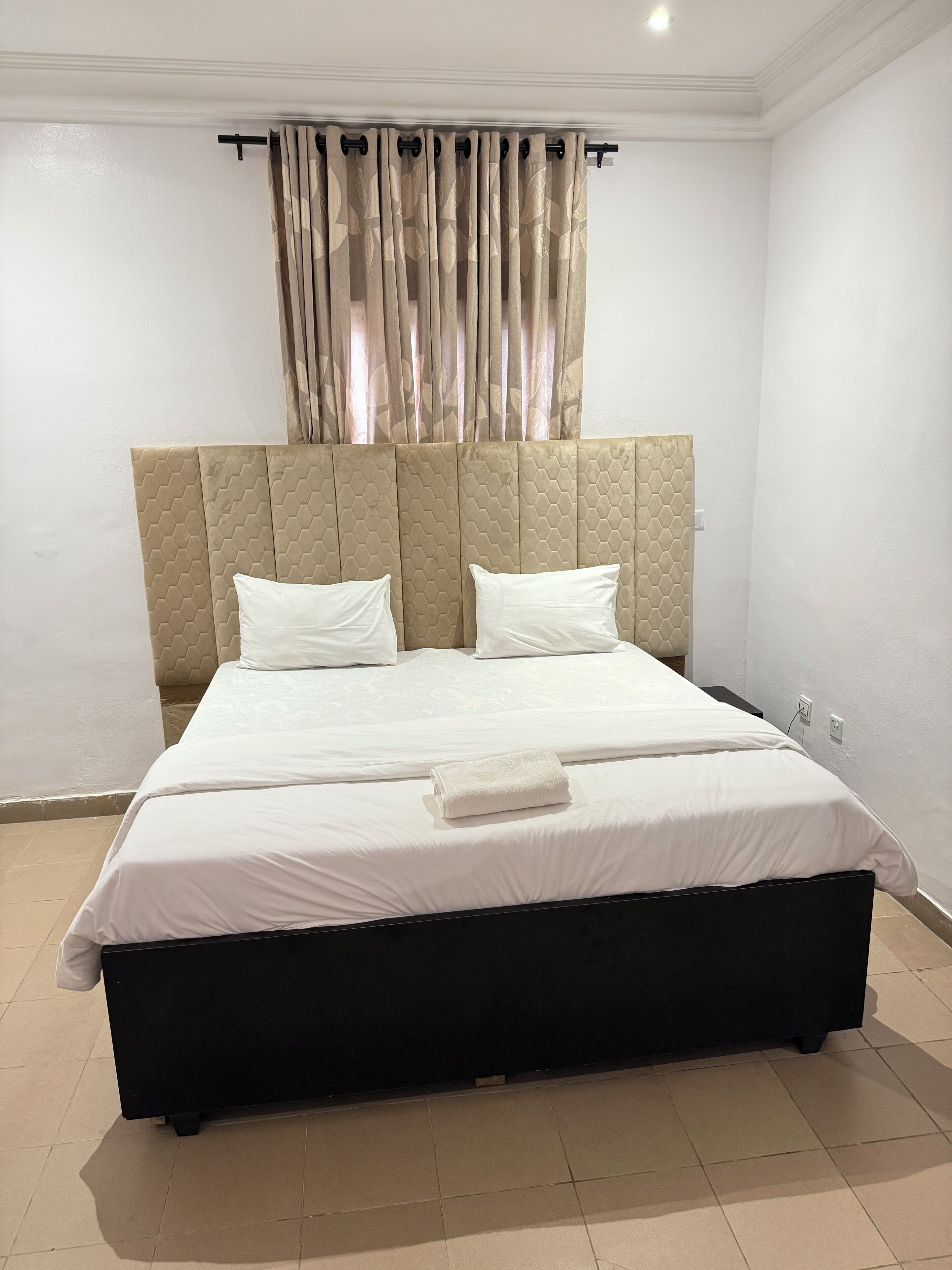 Ein ordentlich bezogenes Bett mit weißer Decke und zwei Kissen, ein Kopfteil mit geometrischem Muster und ein gefaltetes Handtuch auf dem Bett.