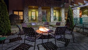 An outdoor patio with a fire pit surrounded by chairs, a stone building with green railings in the background, and a well-lit entrance. - Cozy Mountain Escape at Wyn Smoky Mountains -  1 Bd. – Family Fun Near Dollywood (Sevierville)