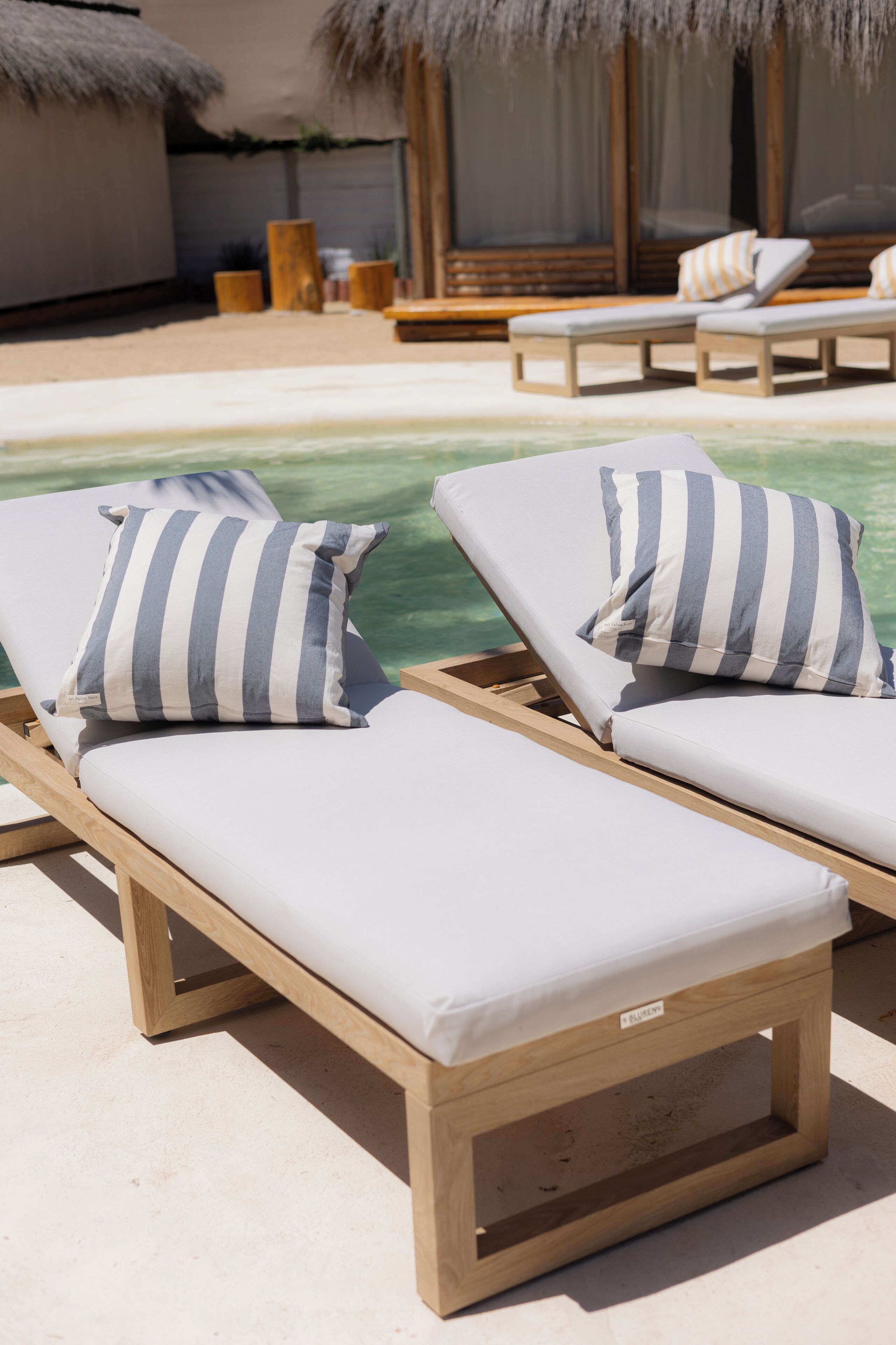 Deux fauteuils de jardin en bois, agrémentés de coussins à rayures, sont placés au bord d’une piscine.