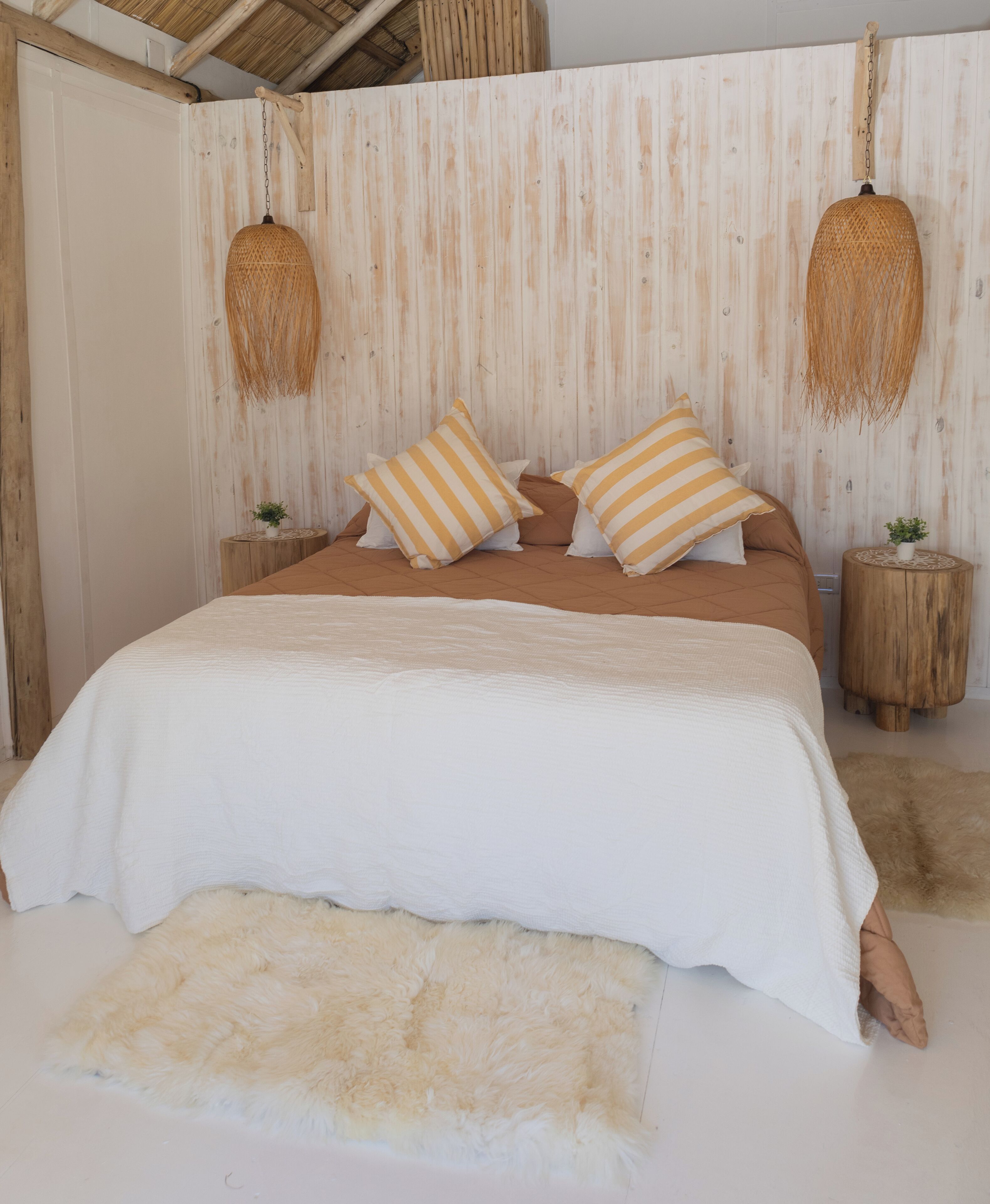 Un lit recouvert d’une courtepointe blanche, avec des oreillers marron et un tapis blanc et moelleux. On y trouve deux tables de chevet en bois, sur lesquelles sont posées de petites plantes en pot.