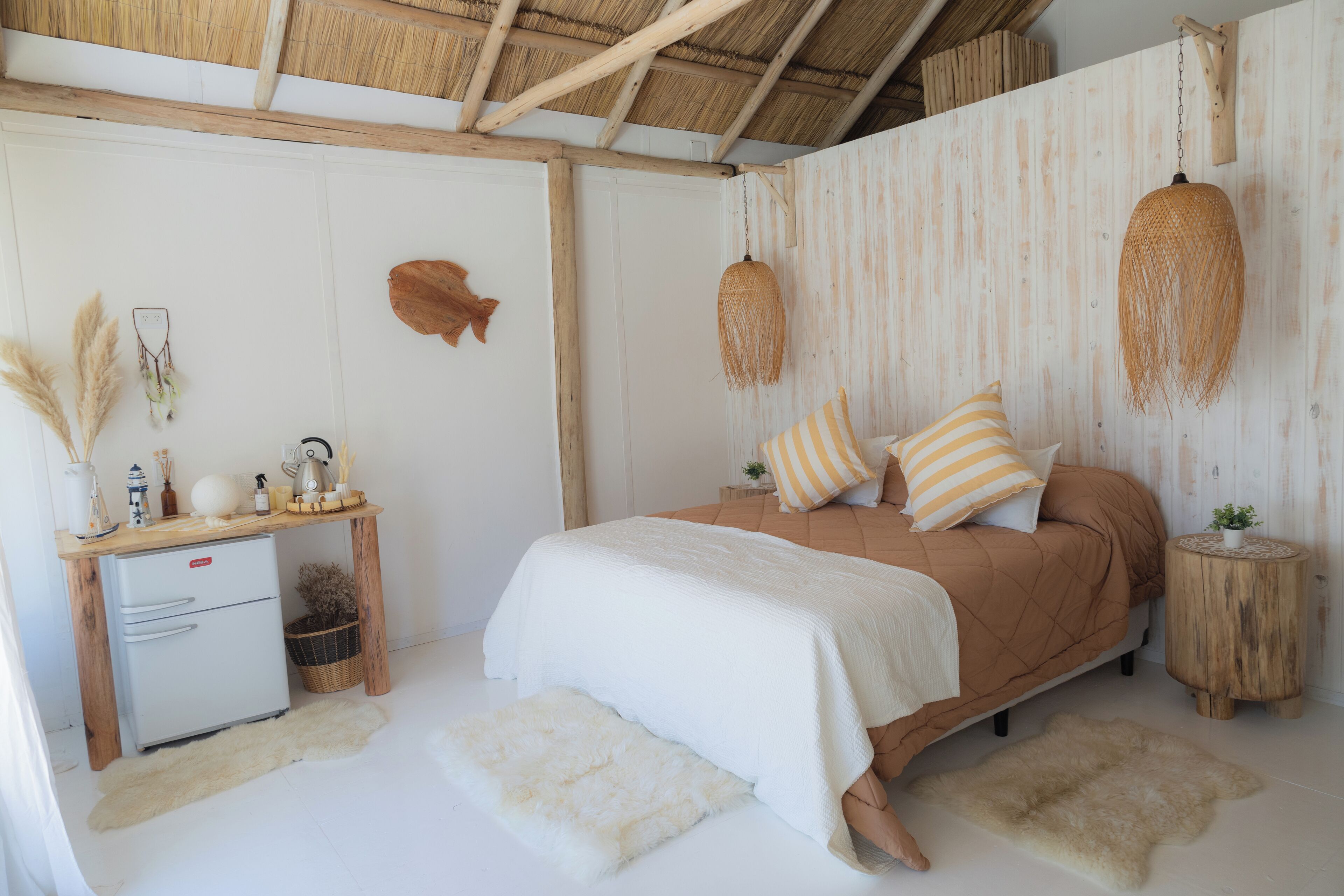 A bedroom with a bed, a small desk, a refrigerator, and a hanging light fixture.