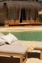 Una cabaña con techo de paja, piscina, terraza de madera y sillones de descanso.
