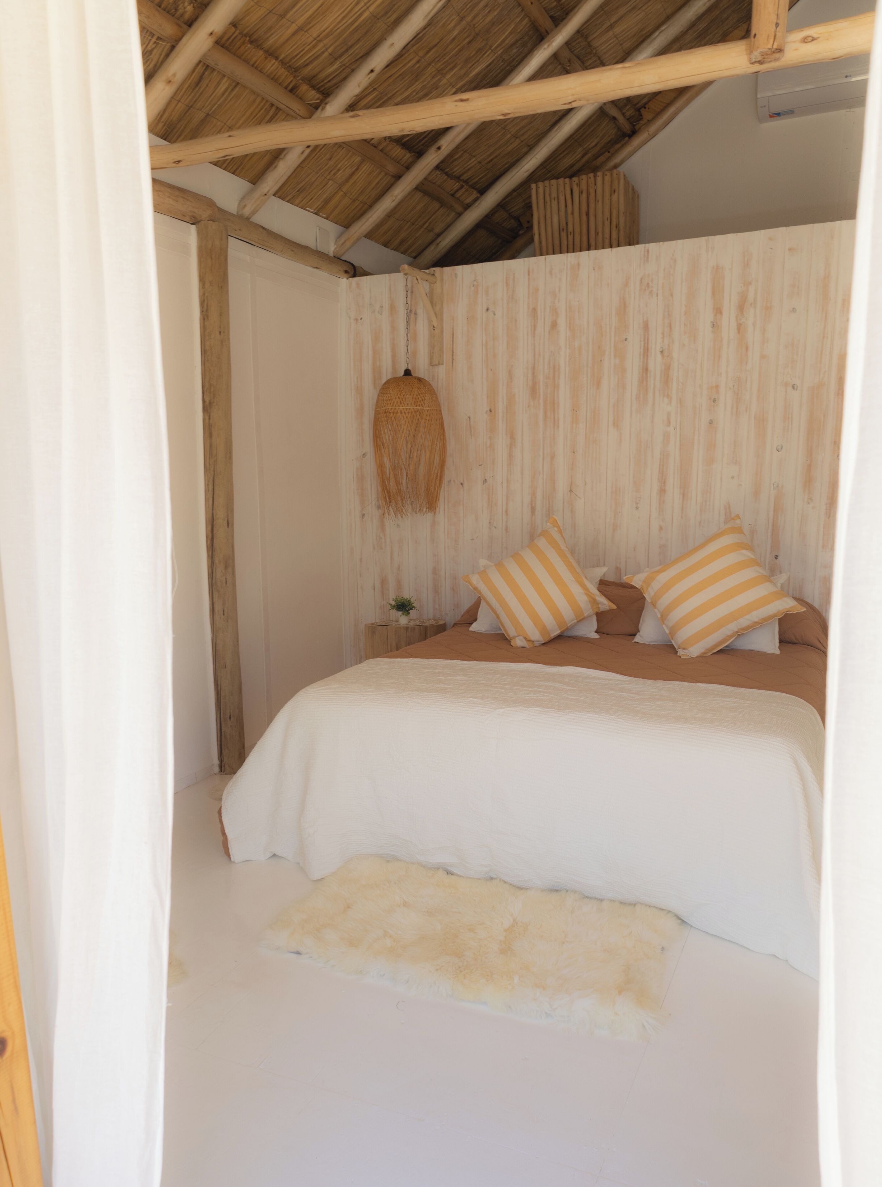Un dormitorio con techo de madera, una cama con ropa de cama blanca y almohadas a rayas, una mesita de noche con una planta y una alfombra blanca y mullida.