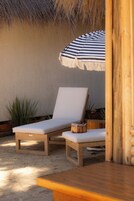 Una silla de lounge de madera con cojín blanco, una sombrilla de playa a rayas y una pequeña mesa de madera.