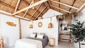 A bedroom with a thatched roof, a bed with a white and gray quilt, a wooden side table, a woven basket, and a plant. - Tribu Eco Beach (Francisco Alvarez)