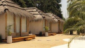 A row of thatched-roof cabins with wooden benches and potted plants, situated on a sandy beach with palm trees. - Tribu Eco Beach (Francisco Alvarez)