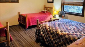 Una habitación con dos camas, una con cubrecama rojo y la otra con cubrecama acolchado. Hay una silla de madera y una mesita con una lámpara.