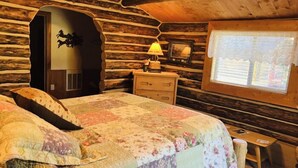 A cozy log cabin room with a bed, a nightstand, a lamp, and a window with a view.