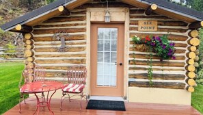 Une cabane en rondins avec une enseigne indiquant « SIX POINT » au-dessus de la porte, un petit porche avec une table et des chaises, et un arrangement floral sur le côté droit.