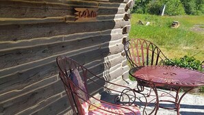 Exterior d'una cabana rústica de fusta amb una taula i cadires metàl·liques, un rètol de fusta i un jardí amb gespa.