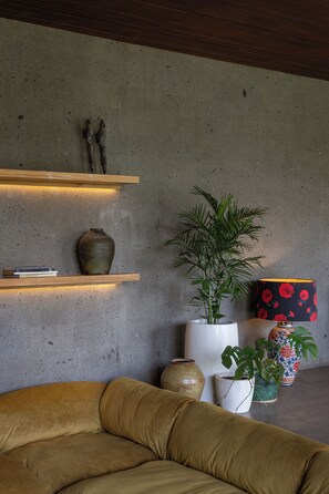 Ein modernes Wohnzimmer mit einer Betonwand, einem Holzregal mit Büchern und einer Vase, einer Couch und Topfpflanzen.