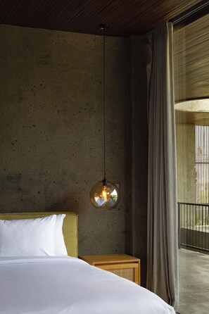 Ein Schlafzimmer mit einem großen Bett, einer hängenden Leuchte, einem Nachttisch aus Holz und Blick durch ein Fenster mit Vorhängen.