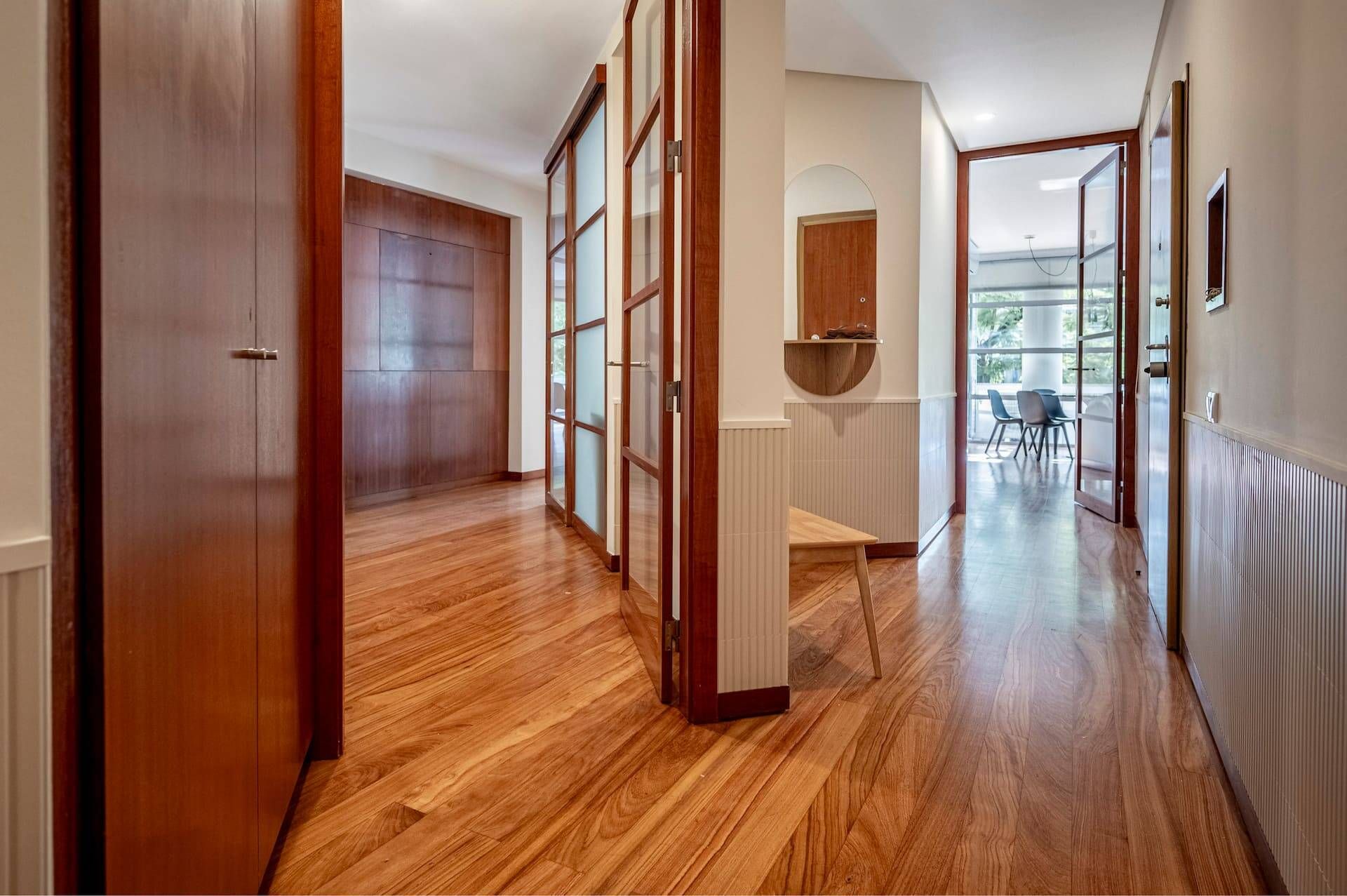 A hallway with wooden flooring, a wooden door, and a view into a dining area with chairs and a table.
