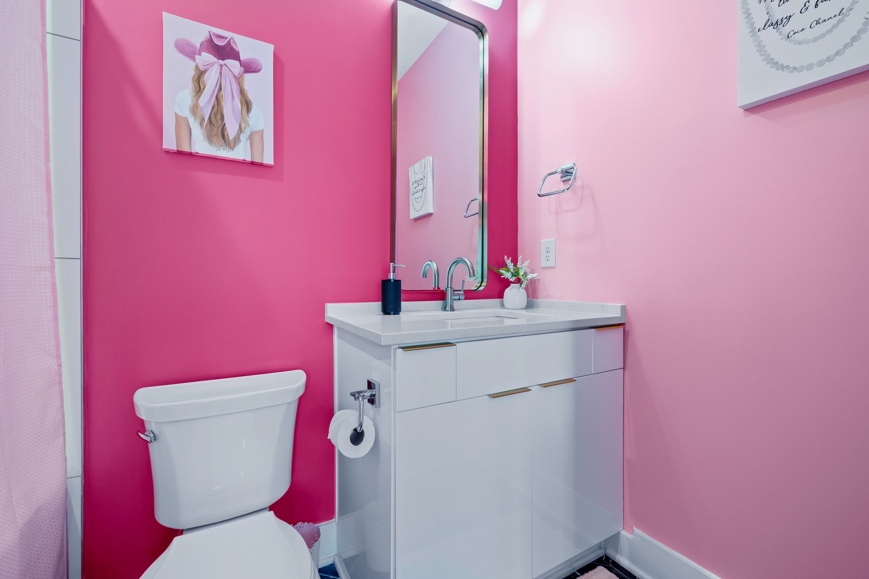 Un baño con pared rosa, inodoro blanco, lavamanos con espejo y un cuadro enmarcado en la pared.