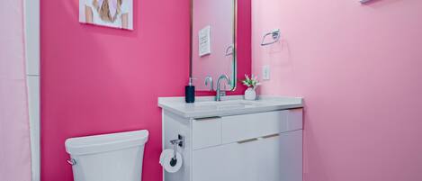Un baño con pared rosa, inodoro blanco, lavamanos con espejo y un cuadro enmarcado en la pared.