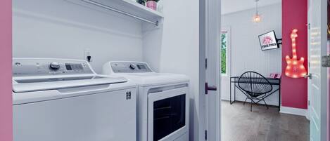 A laundry room with two washing machines, a small shelf with a plant, a chair, and a guitar-shaped light fixture.