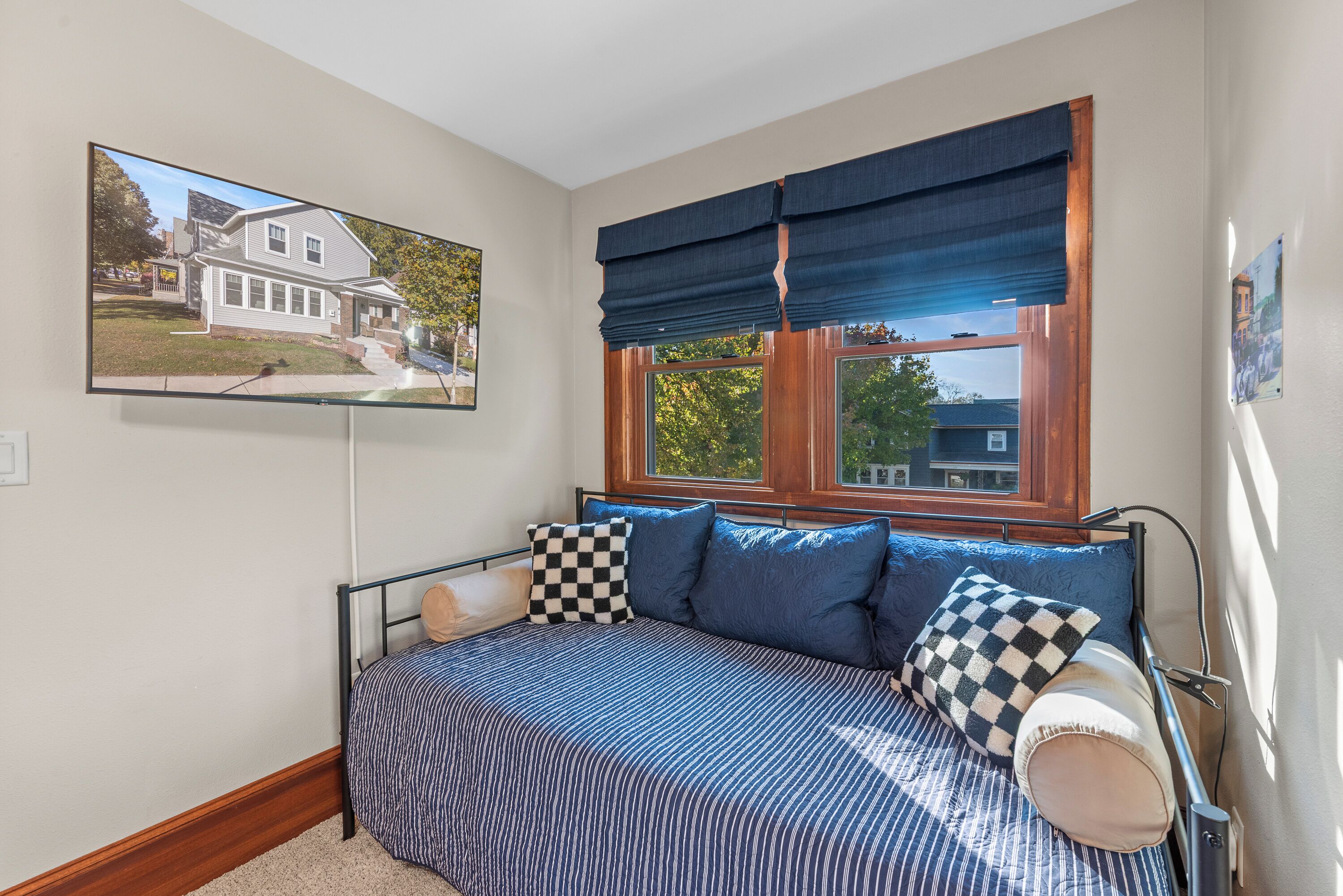 Un dormitorio con una cama, un ventanal con cortina azul, un televisor en la pared y un cuadro con una imagen de una casa.