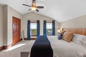 Une chambre avec un grand lit, un ventilateur de plafond et deux fenêtres avec des rideaux bleus.